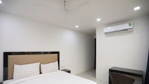 A modern hotel room with a large bed, a desk, a chair, a ceiling fan, and an air conditioning unit. - Hotel Raaso Inn Tuglkabad (New Delhi)