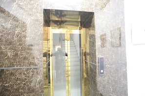 An elevator with a marble wall and a glass door. - Hotel Raaso Inn Tuglkabad (New Delhi)