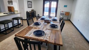 A dining area with a long wooden table set for six, surrounded by black chairs. Above the table, there are three framed abstract paintings.