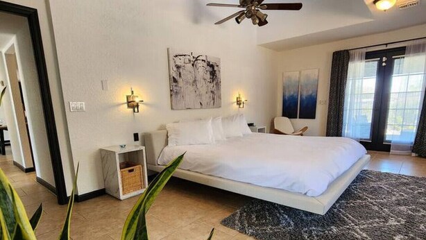 A modern bedroom with a large bed, ceiling fan, artwork on the wall, and a view of the outdoors through the window.