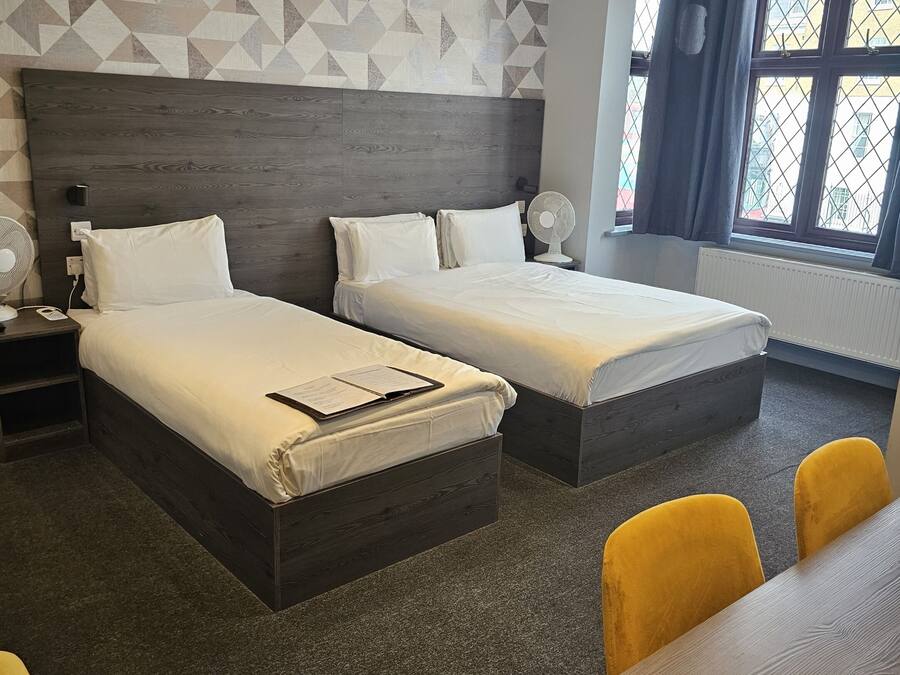 Habitación de hotel con dos camas, un escritorio, sillas, un ventilador de pared y una ventana con cortinas.