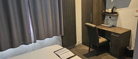 A hotel room with a bed, a desk, a chair, a TV, and a small shelf with a kettle and glasses.