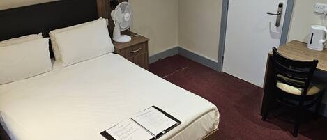 A hotel room with a bed, bedside table with a fan, a desk with a chair, a red hanging light, and a door with a fire evacuation plan.