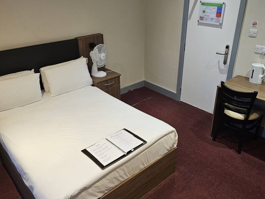 Habitación de hotel con cama, mesita de noche con ventilador, escritorio con silla, lámpara colgante roja y puerta con plan de evacuación contra incendios.