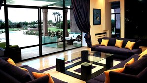 A modern living room with a glass coffee table, dark sofas with yellow cushions, and a view of a garden through large windows. - VICTOR HOME RENT (Jbilate)