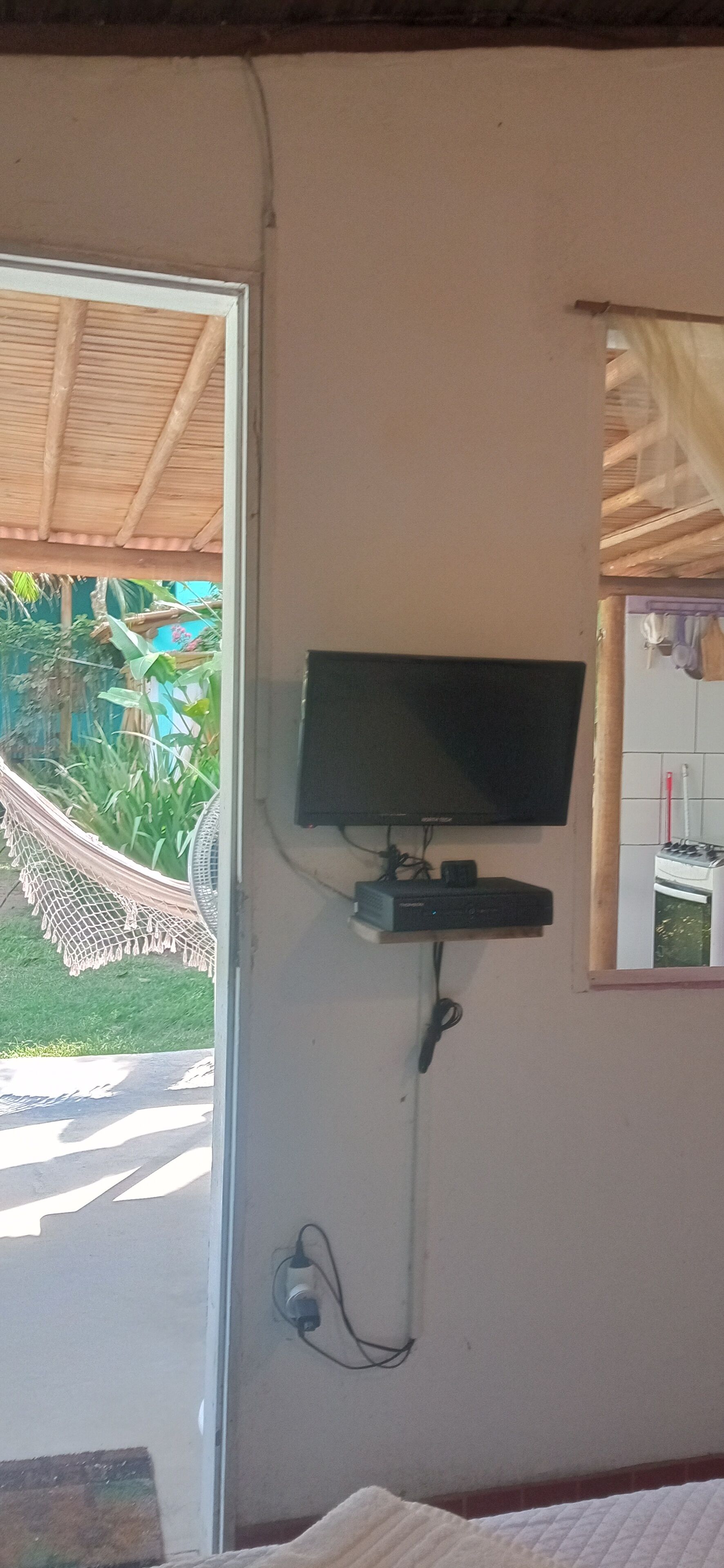 A room with a television mounted on the wall, a hammock outside the window, and a small kitchen area visible through the window.