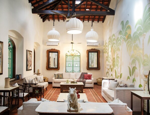 A spacious living room with a high ceiling, large windows, and a mural of palm trees. - Lamrin Ucassaim Goa (Bastora)