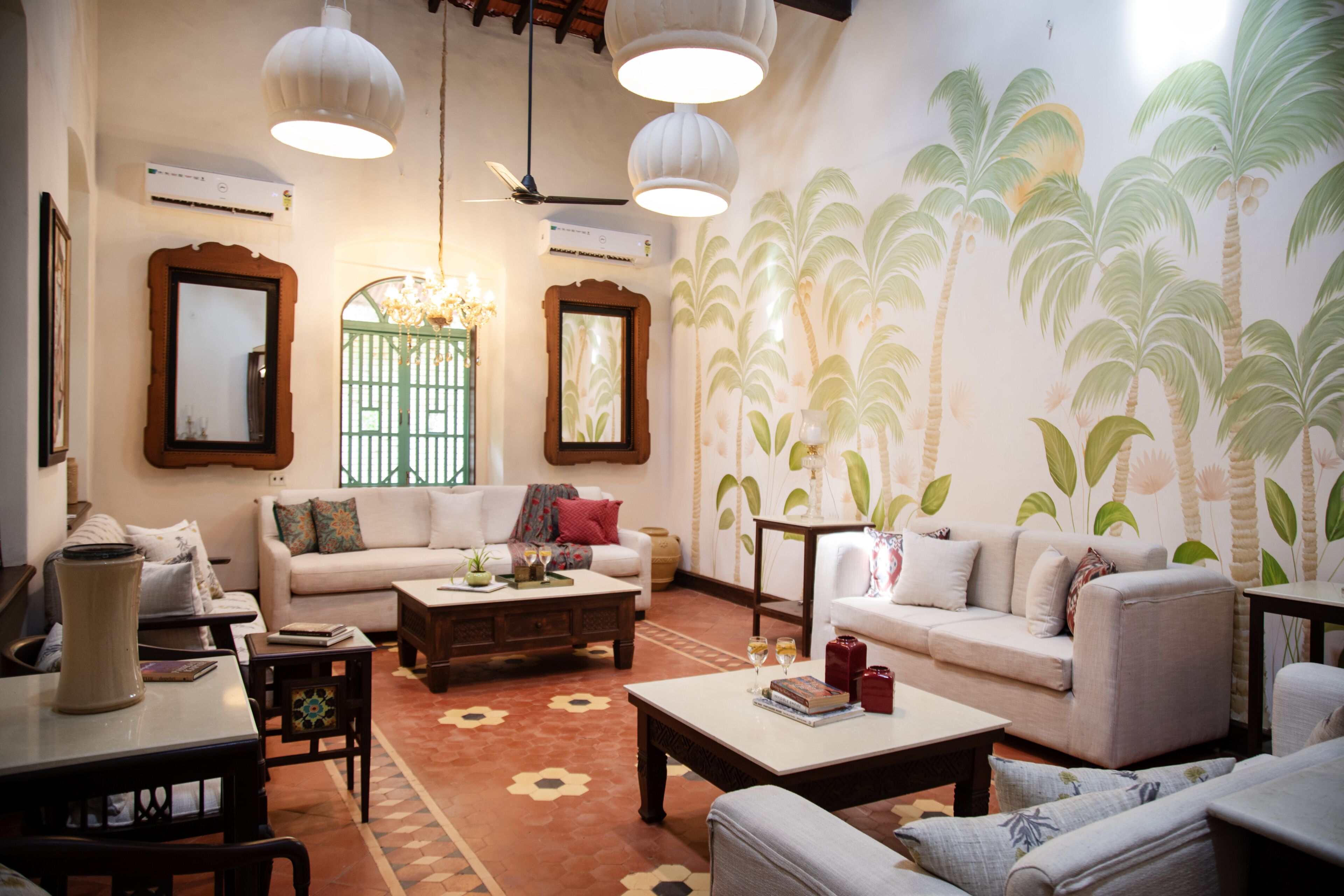 Un salón espacioso con un sofá grande, una mesa de centro y un tapete estampado. Hay dos ventanas amplias con marcos decorativos y un mural en la pared con palmeras.
