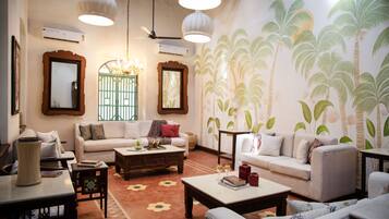 Un salón espacioso con un sofá grande, una mesa de centro y un tapete estampado. Hay dos ventanas amplias con marcos decorativos y un mural en la pared con palmeras.