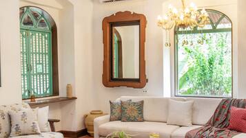 A living room with a sofa, coffee table, and decorative items. There is a window with a view of greenery and a mirror on the wall.
