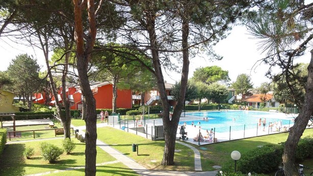 Una piscina rodeada de árboles y una zona verde, con un edificio rojo al fondo.