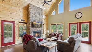 A spacious living room with a stone fireplace, two sofas, a coffee table, and large windows offering a view of the outdoors. - Brand New 5 Bedroom with Arcade, WiFi, Hot Tub!  Located in Thunder Mtn Resort! (Sevierville)