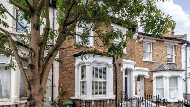 A row of terraced houses with white-framed windows, a tree in the foreground, and a black metal fence. - 5 Bed Apartment in Hammersmith (London)