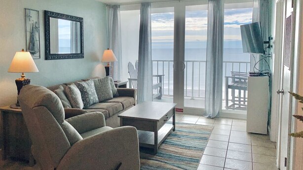 A living room with a sofa, a coffee table, and a TV. There are two lamps, a rug, and a view of the ocean through large windows.