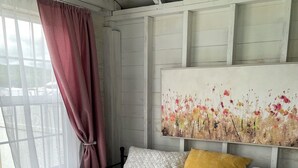 A bedroom with a bed, a window with curtains, a painting on the wall, and a wooden ceiling.
