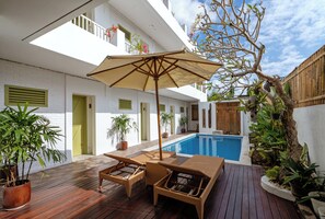 Una terraza de madera con piscina, muebles de mimbre y plantas en macetas.