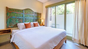 A neatly made bed with white linens and orange pillows, a wooden headboard with intricate carvings, a bedside table with a lamp, and a large window with sheer curtains. - Honu Studio Living Bali (Pererenan)