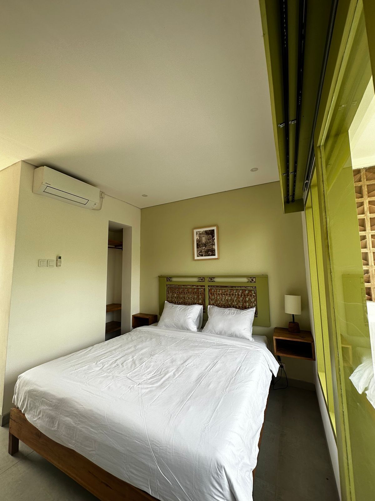 A hotel room with a bed, a nightstand, a wall-mounted air conditioner, a framed picture, and a window with blinds.