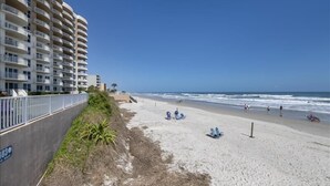 A beach with a high-rise building in the background, a sandy shore, and a few people and chairs. - 705 St Maarten - Daytona Beach Oceanfront Vacations, Inc (Daytona Beach Shores)