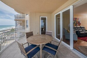 Un balcon avec une table ronde en bois et des chaises, une vue sur la plage, et un espace de vie comprenant un canapé et un tableau.