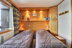 A room with two beds, wooden walls, a window with curtains, a wall-mounted shelf with a lamp, and a wooden cabinet.