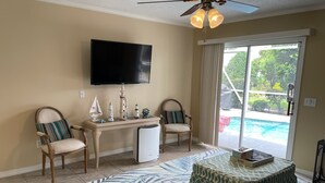 A living room with a flat-screen TV, two chairs, a coffee table, and a view of a swimming pool through a sliding glass door. - Welcome to Your Ideal Getaway in The Villages! (The Villages)