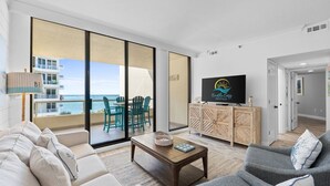 Una sala de estar moderna con un sofá, un sillón y una mesa de centro. Hay un televisor sobre un mueble de madera, y un amplio ventanal corredera que da a un balcón con vistas al océano.
