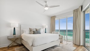 A bedroom with a large bed, a ceiling fan, a nightstand with a lamp, and a view of the ocean through large windows.