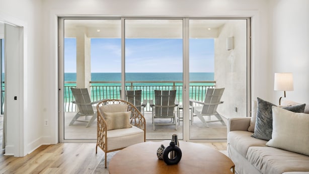 A spacious room with a large sliding glass door leading to a balcony with a view of the ocean. There are several chairs and a sofa on the balcony, and a table with a vase and a lamp inside the room.