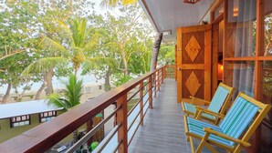 Un balcon en bois offrant une vue sur la plage et les palmiers. On y trouve deux chaises à rayures et une table avec des bouteilles.