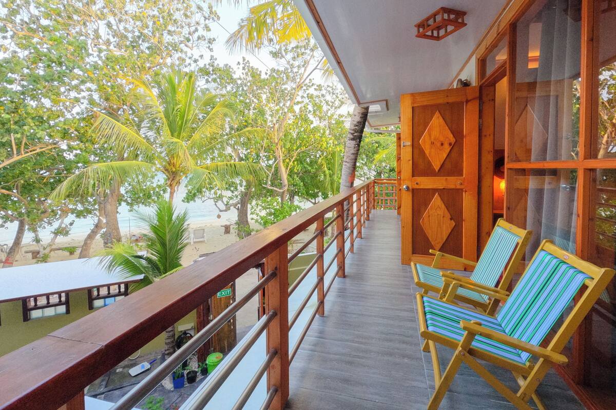 A wooden balcony with a view of the beach and palm trees. There are two striped chairs and a table with bottles.