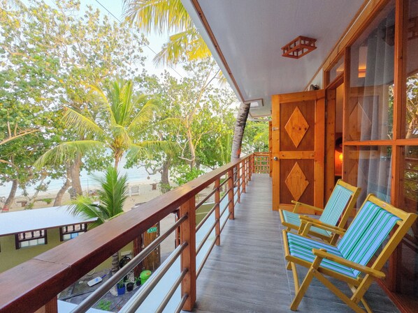 A wooden balcony with a view of the beach and palm trees. There are two striped chairs and a table with bottles. - Dhamana Beach (Mahibadhoo)