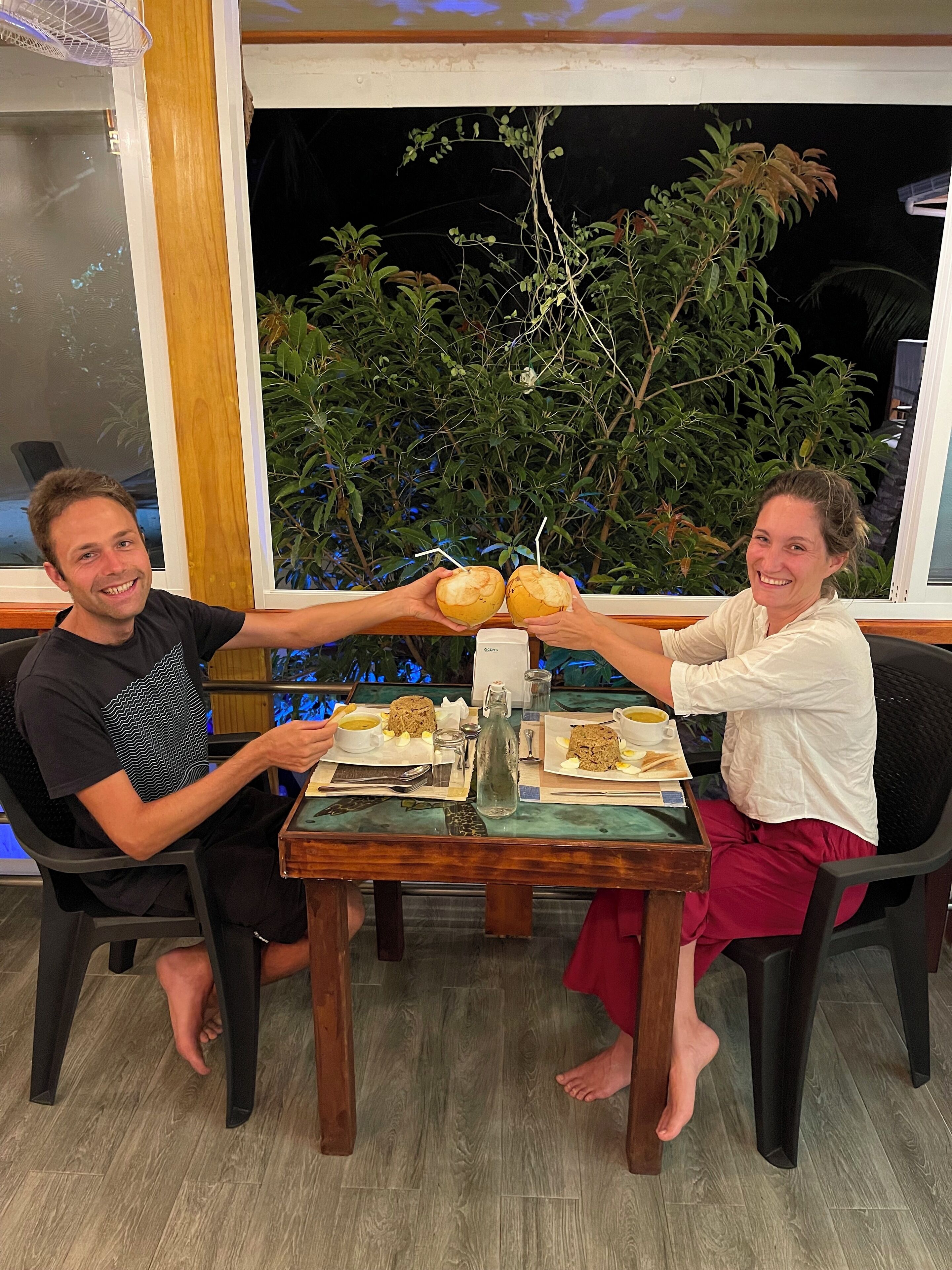 Deux personnes sont assises à une table, devant un repas, avec une fenêtre et une plante en arrière-plan.