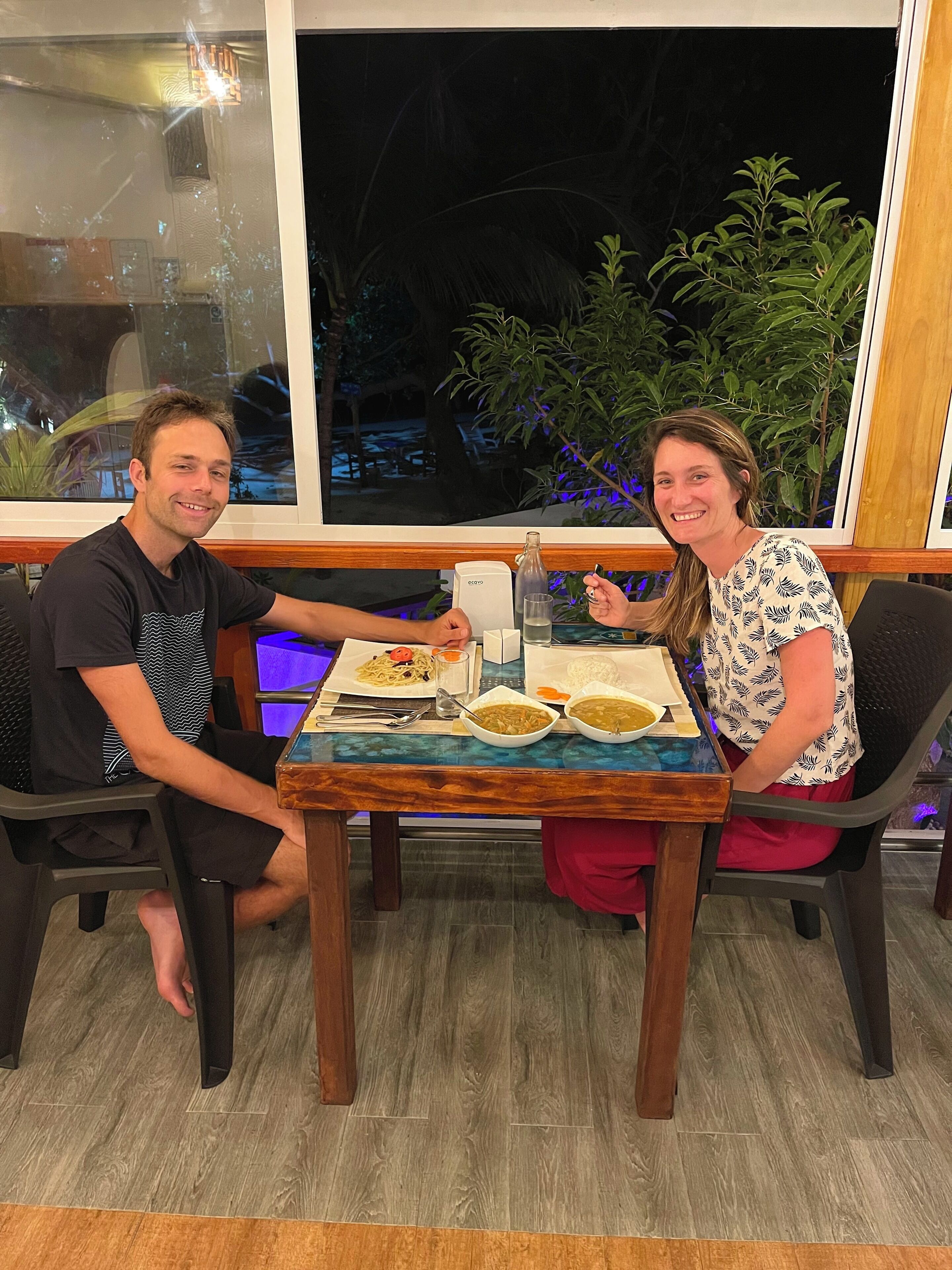 Two people seated at a table with a meal, a window with a view of greenery, and a wooden floor.