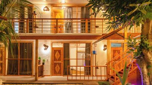 Une maison en bois à deux étages, dotée d’un balcon, de grandes portes coulissantes et d’un petit jardin avec une terrasse en bois et un élément décoratif aquatique.