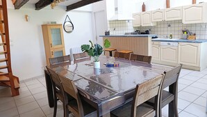 Un coin repas avec une grande table et des chaises en bois, une plante en pot et une cuisine en arrière-plan.