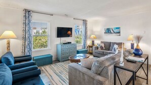 A modern living room with a sofa, armchairs, a coffee table, and a TV. There are curtains, a painting, and a lamp on a side table. - Pleasant Seaprise: Private Pool, 200ft. to Beach (Nags Head)