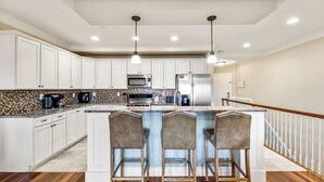 A modern kitchen with white cabinets, a central island with bar stools, stainless steel appliances, and a patterned backsplash. - Surfing Gecko: Oceanfront, Shared Pool, Dogs OK (Kill Devil Hills)