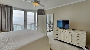 Una camera d'albergo con un letto, una televisione su un comò, una ventola a soffitto e vista sull'oceano attraverso ampie finestre.