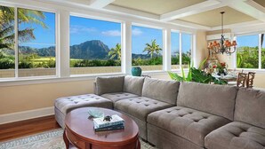 A living room with a sectional sofa, a round coffee table, and large windows offering a view of a mountain and palm trees. - Pinnacle at Poipu -  6 Bedroom Luxury Estate - Amazing Views & Salt Water Pool! (Koloa)