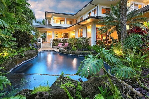 A pool surrounded by lush greenery, a covered patio with seating, and a two-story building with large windows.