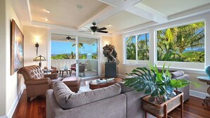 A spacious living room with a large gray sofa, a coffee table, and a potted plant. There is a sliding glass door leading to a balcony with a view of palm trees. - Pinnacle at Poipu -  6 Bedroom Luxury Estate - Amazing Views & Salt Water Pool! (Koloa)