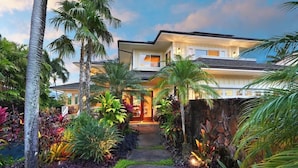 A modern two-story house with a stone pathway leading to the entrance, surrounded by lush tropical plants and palm trees. - Pinnacle at Poipu -  6 Bedroom Luxury Estate - Amazing Views & Salt Water Pool! (Koloa)