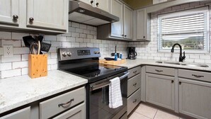 Une cuisine avec des meubles blancs, une hotte en acier inoxydable, une cuisinière noire et une fenêtre avec des stores.