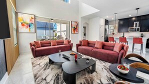 Un vaste espace de vie comprenant un grand canapé d’angle, une table basse et une table à manger avec des chaises. Au fond, on trouve une cuisine équipée d’appareils électroménagers modernes et dotée de tabourets de bar.