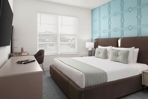 Ett hotellrum med en säng, ett skrivbord, en stol, en tv och ett fönster med persienner.