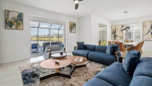 Ein geräumiges Wohnzimmer mit einem großen, blauen Sofa, einem Couchtisch und einem Teppich. Es gibt einen Essbereich mit Stühlen und einem Tisch. Das Zimmer verfügt über große Fenster und Kunstwerke an den Wänden.