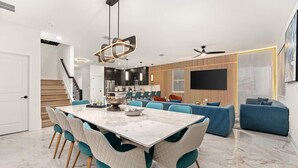 A modern kitchen with dark cabinetry, a central island, and a dining area with a marble table and teal chairs.