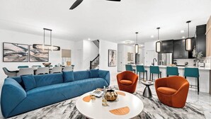 A modern living room with a blue sectional sofa, two orange armchairs, a round coffee table, and a kitchen area with bar stools.