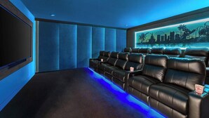 A home theater room with a row of black leather recliner seats, a large flat-screen TV, and a cityscape mural on the wall. - Magnificent Mansion | Breathtaking estate mins to Universal, Disney, restaurants (Orlando)